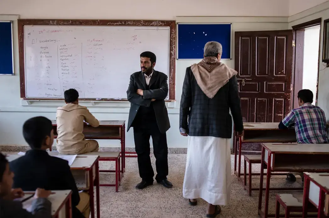 Hussein al Tawushi (center left), principal at the school where Yahya (center right) teaches, looks over students as they take their final exams. Image by Alex Potter. Yemen, 2018.
