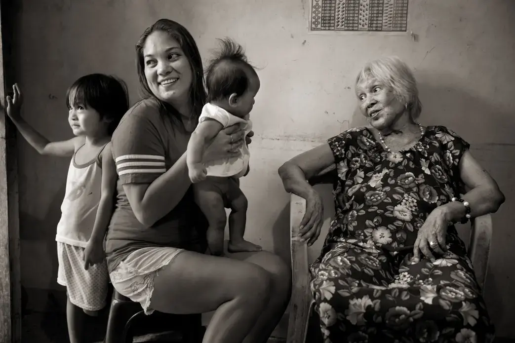 After singing for soldiers, Felicidad delos Reyes (right) was selected to 'receive a gift' at the garrison behind her school. She was locked in a room; later five soldiers jumped on her, held her down and beat her into submission as they took turns raping her. Here, she is photographed with her great-granddaughter Lia Maglanit, great-grandson Elijah Maglanit and granddaughter Jonalyn Patiman. Image by Cheryl Diaz Meyer. Philippines, 2019.