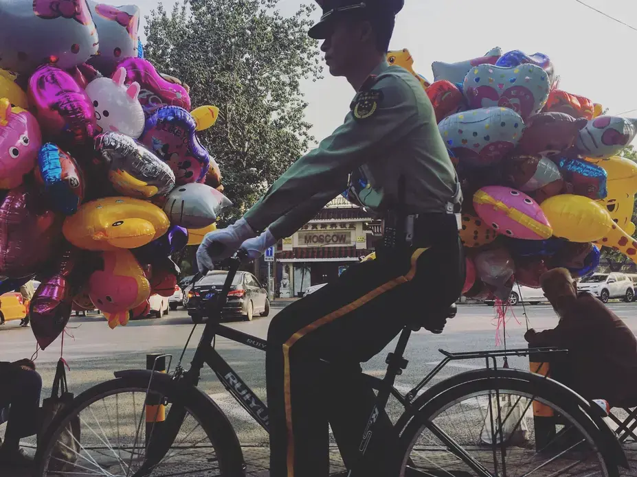 A member of the Chinese military rides his bike past two men selling balloons on Sunday, Sept. 24, 2017, on the same block as the residence of U.S. Ambassador to China Terry Branstad in Beijing, China. Image by Kelsey Kremer. China, 2017.