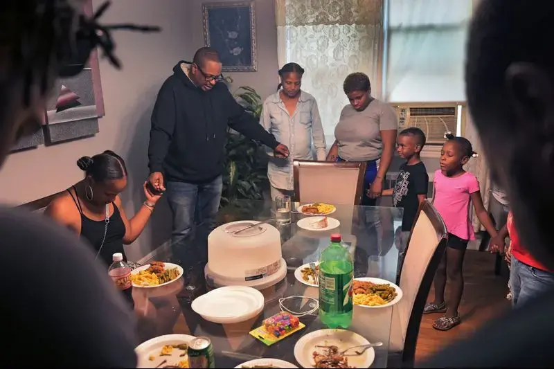 Cecil Burton – viewed as a 'deadbeat dad' by many for his child support debt – leads his family in prayer: 'Father God, thank you for showing my family what true love is. Let’s eat and have fun and enjoy ourselves.' At 54, much of the debt he owes is to the state to repay welfare benefits his children received when they were young. Image by Karl Merton Ferron / Baltimore Sun. United States, 2019. 
