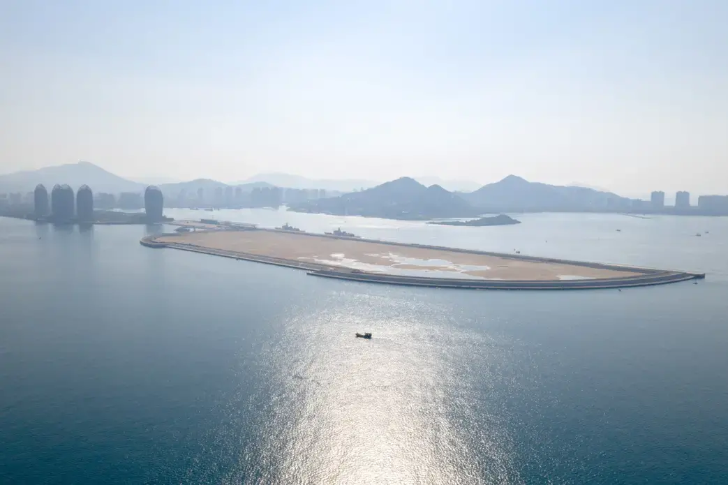 A view from on Phoenix island, Sanya, Hainan. Phoenix Island, on the resort city of Sanya, was the Hainan island’s first flagship land reclamation mega project, attracting internationally renowned architects. Reclaimed between 2002 and 2003, and opened for business in 2015, the artificial island in Sanya Bay has luxury apartments, hotels and a cruise centre—all a short distance from the centre of the city. A second phase of reclamation was undertaken in 2014—seen here as the empty, sandy plot—bringing the total size of the island to 0.84 square kilometers. But this was found to have led to the erosion of over 2km of Sanya Bay’s coast. Following an environmental inspection in late 2017, the government has slapped a suspension on further construction as the project was found to have impacted coastal erosion and the ecosystem, and caused sedimentation at the mouth of the Sanya River. Companies involved in this project include New York-based Balmori Associates, which does landscape and urban design, and the brand-name Chinese architect Ma Yansong's firm MAD. Image by Sim Chi Yin. China, 2018. 