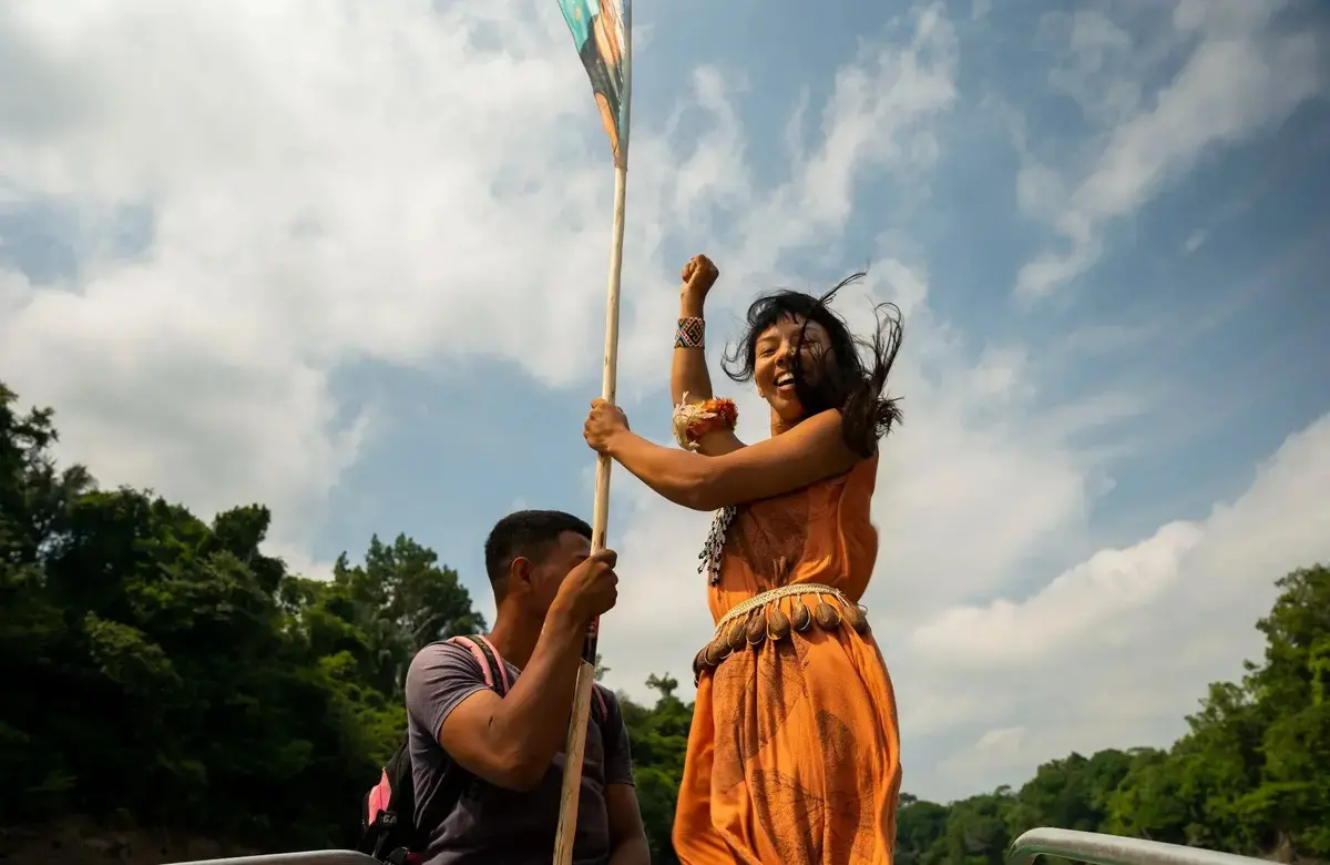 A woman raises her fist triumphantly while holding a flag and standing on a boat. Another person holds the bottom of the flag and looks away from the camera. 