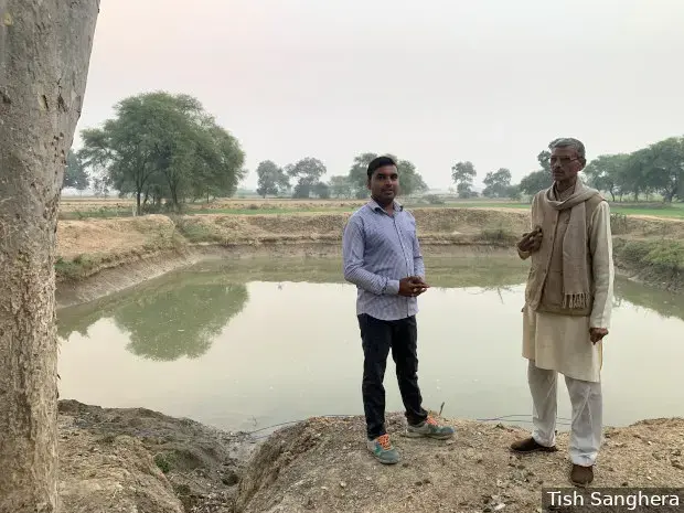 Nawal Kishore Diwedi (on right) with his son-in-law, Vedprakash Diwedi, in front of a pond built under the ‘Khet Talab Yojana’, or ‘Farm Pond Scheme’. They no longer struggle to irrigate their fields and have observed a rise in groundwater levels. Image by Tish Sanghera. India, undated. </p>
<p>