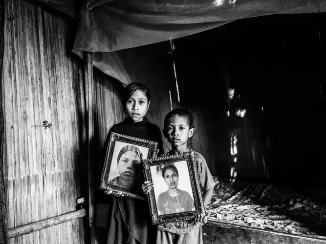Dolfina’s children Erna and Julius hold photos of their mother inside their hut in East Nusa Tenggara, Indonesia. They now live with their grandfather Mikhael. Image by Xyza Bacani. Indonesia, 2018.<br /> 