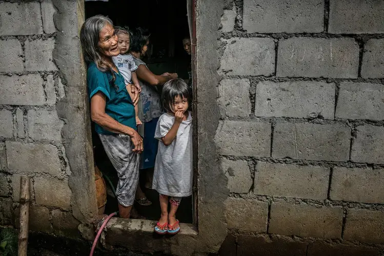Remy Fernandez, 84, holds her grandchild, RJ, in the front door of her cinder block house, built beside a huge rubbish dump in Payatas, a neighborhood in Metro Manila. She is raising seven grandchildren because her son, Constantino de Juan, a drug user, was killed by masked men. The assassins entered the house through this front door. Police deny claims by rights groups that the vigilantes who carry out many such killings are paid by them. Image by James Whitlow Delano. Philippines, 2017.