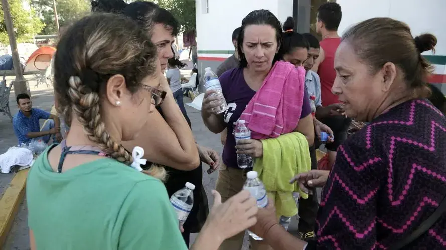 A Mexican volunteer, right, gives water to Cuban migrant women living in a makeshift camp by the Gateway International Bridge in Matamoros, Mexico. Image by Jose A. Iglesias. Mexico, 2019.
