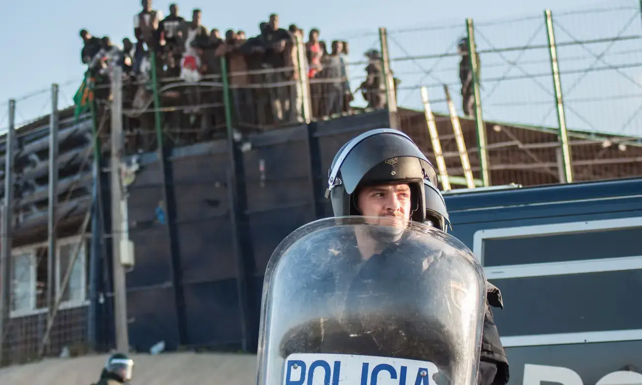 A border agent in riot gear stands in the foreground, with migrants behind a wire fence in the background