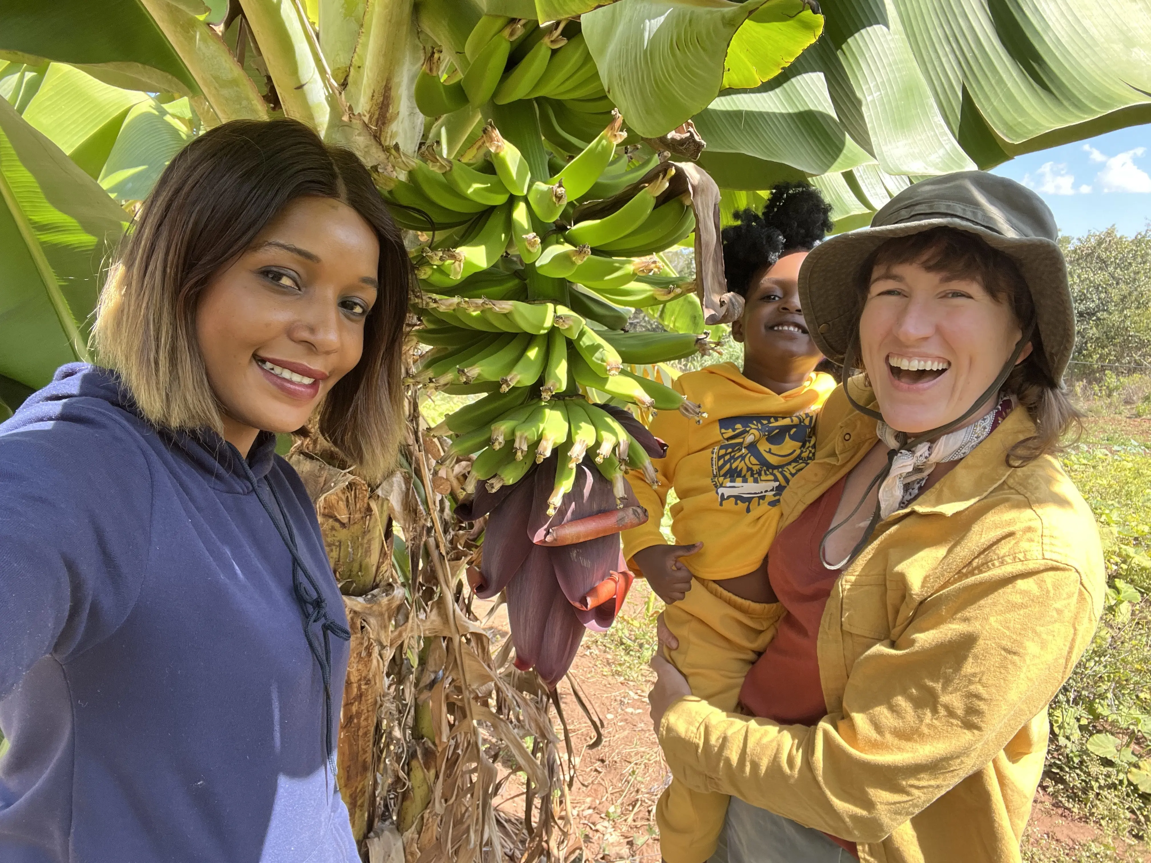 Woman holds a young child while she stands with her friend in front of banana tree