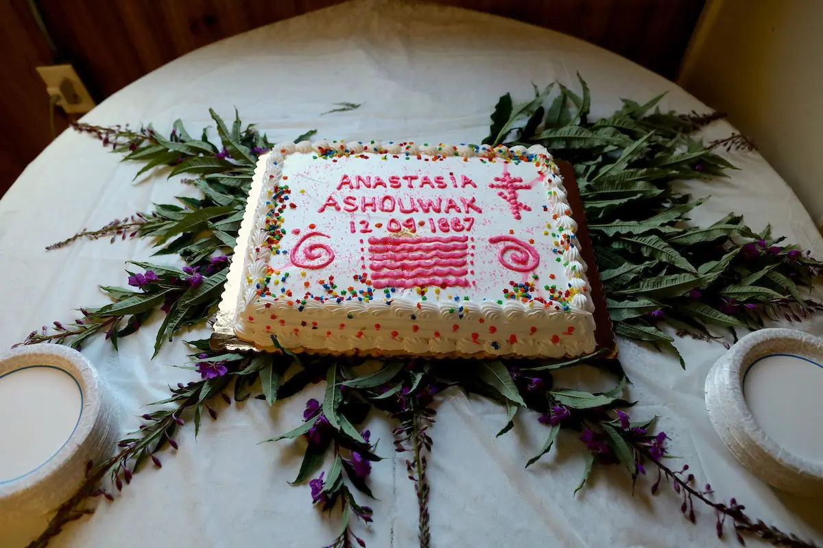 A white cake with rainbow sprinkles sits atop a garland of purple-flowered plants. It reads "Anastasia Ashouwak 12-09-1887" in pink icing.