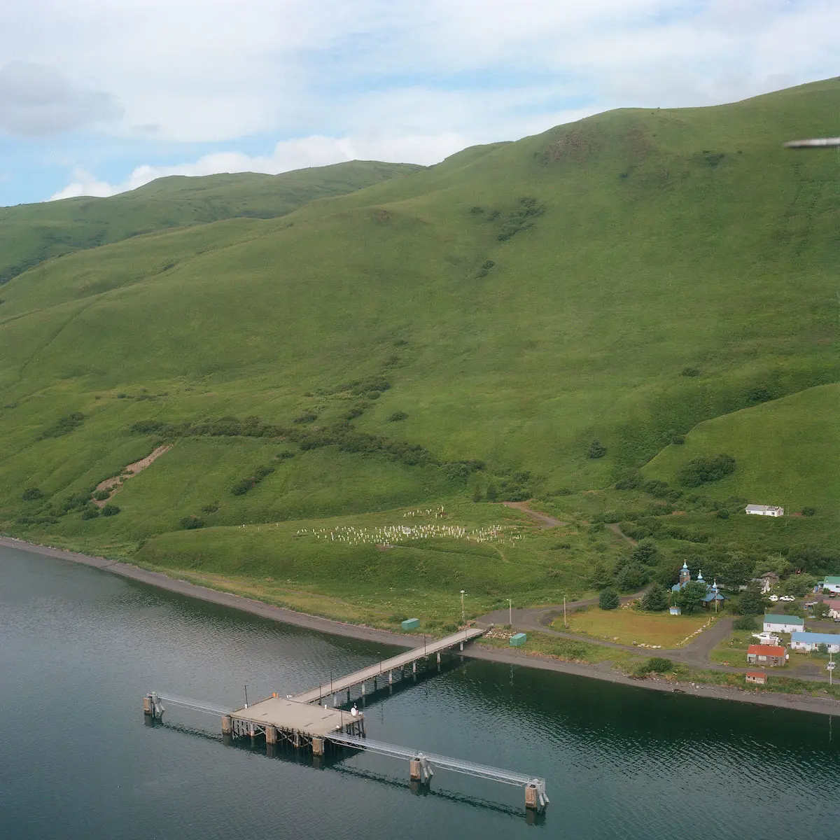 An aerial view of a dock and landing pad over lightly rippled water; it juts out from a lush hillside.