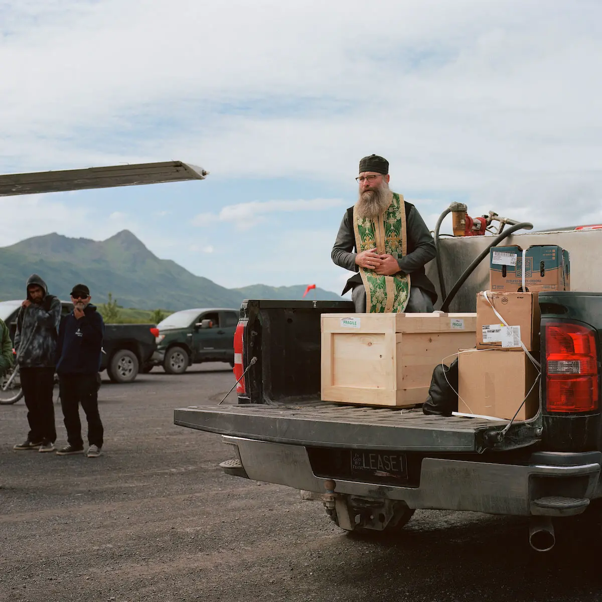 A priest in gilded green robe sits in the back of a pickup with the box containing Anastasia's remains.