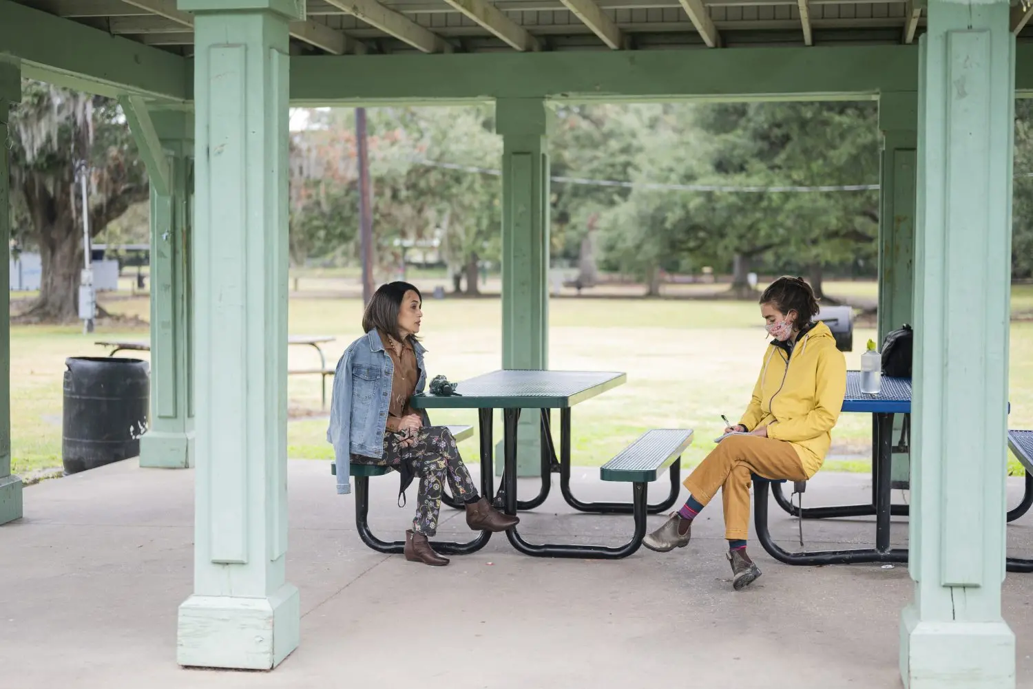 Two people sit socially distanced at picnic tables in a covered patio in a park.