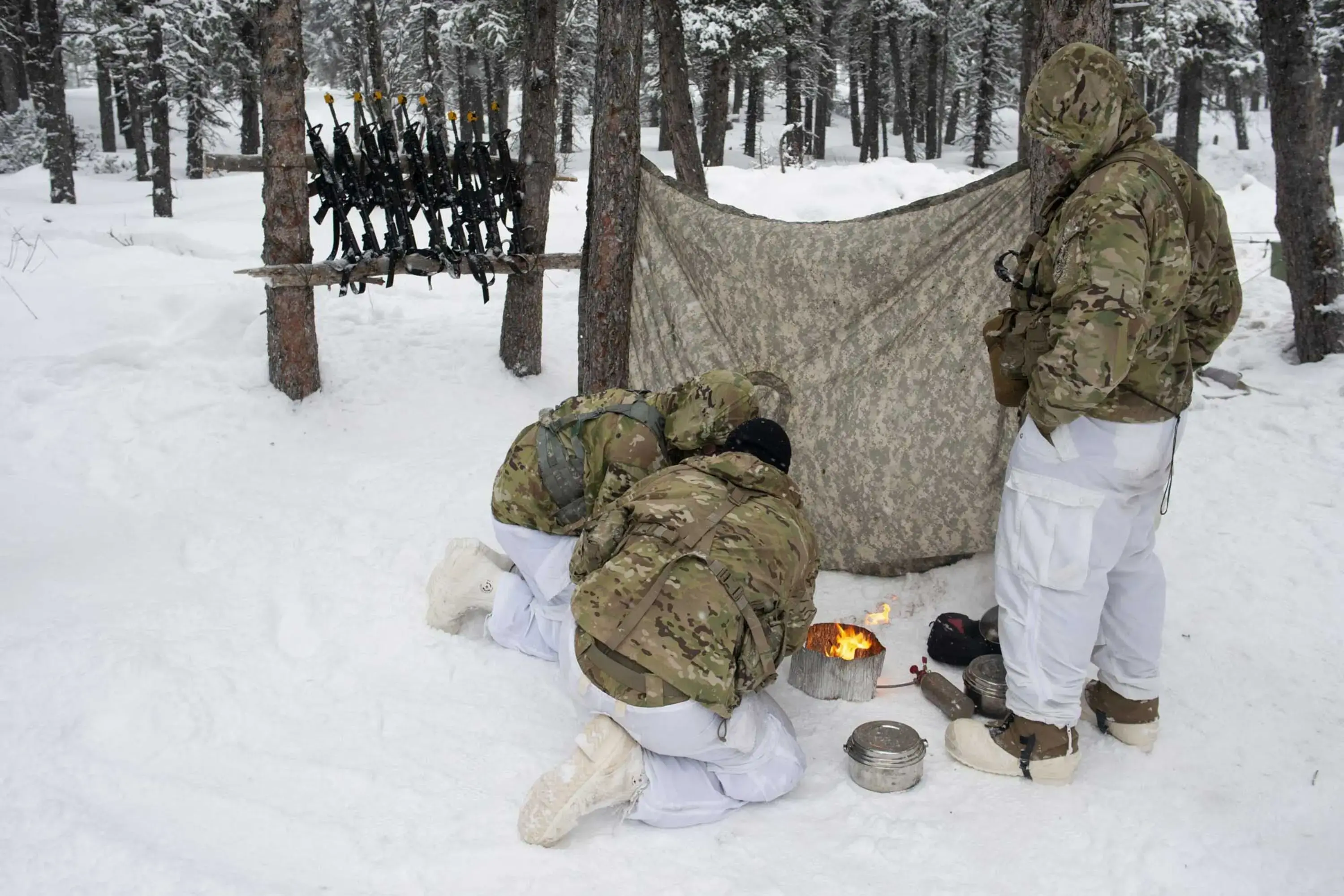 Three soldiers gather around a small stove set up on the snow-covered ground, with their weapons stationed in a row on a rack made from tree trunks.