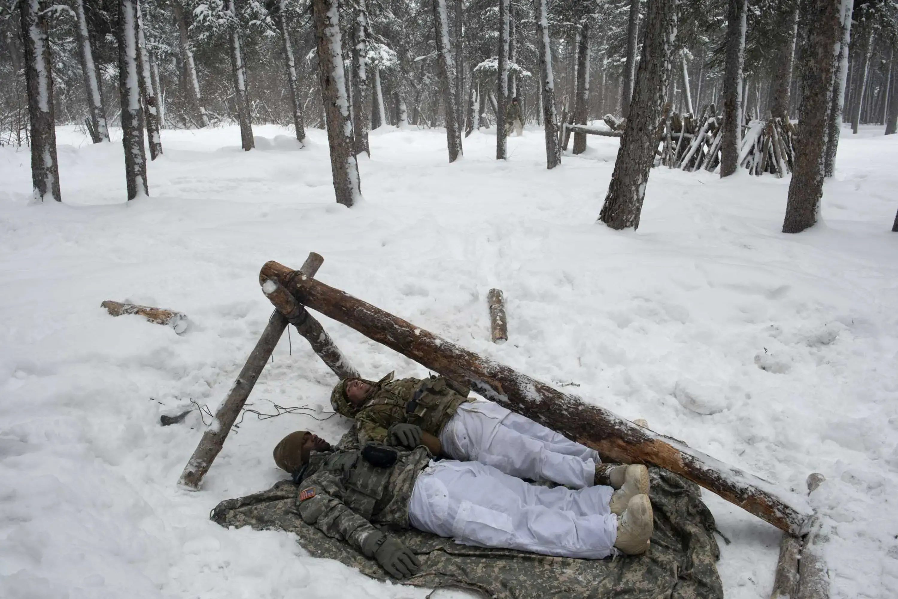Two soldiers lie side by side on a thin sheet upon the snow-caked ground inside a crude wooden frame. They are testing the dimensions of a shelter they are building.