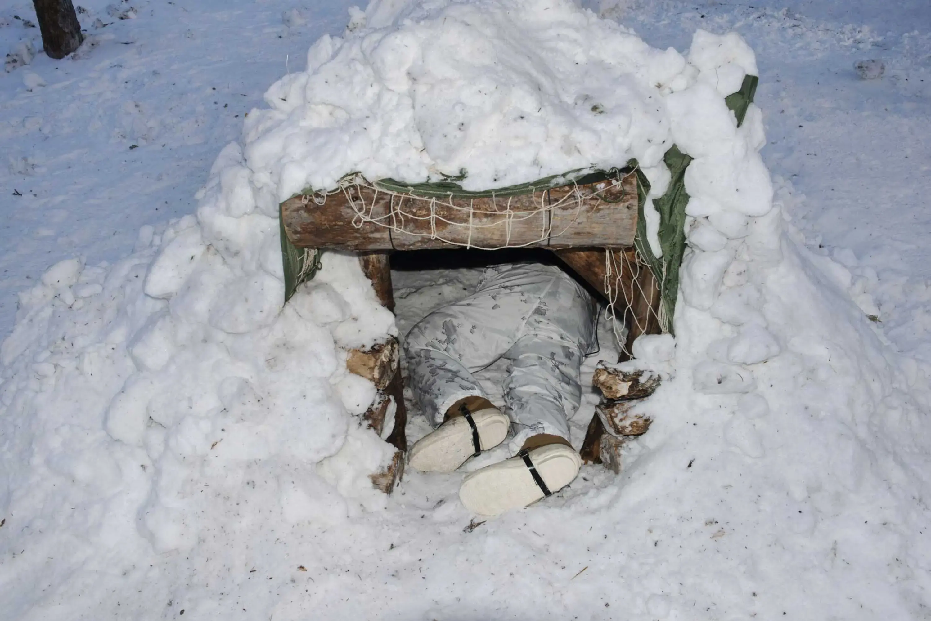 A soldier visible from the waist down is climbing into a shelter that looks like the entrance to a small animal’s burrow. The entrance is framed by wooden trunks and the rest of the shelter is covered in snow.
