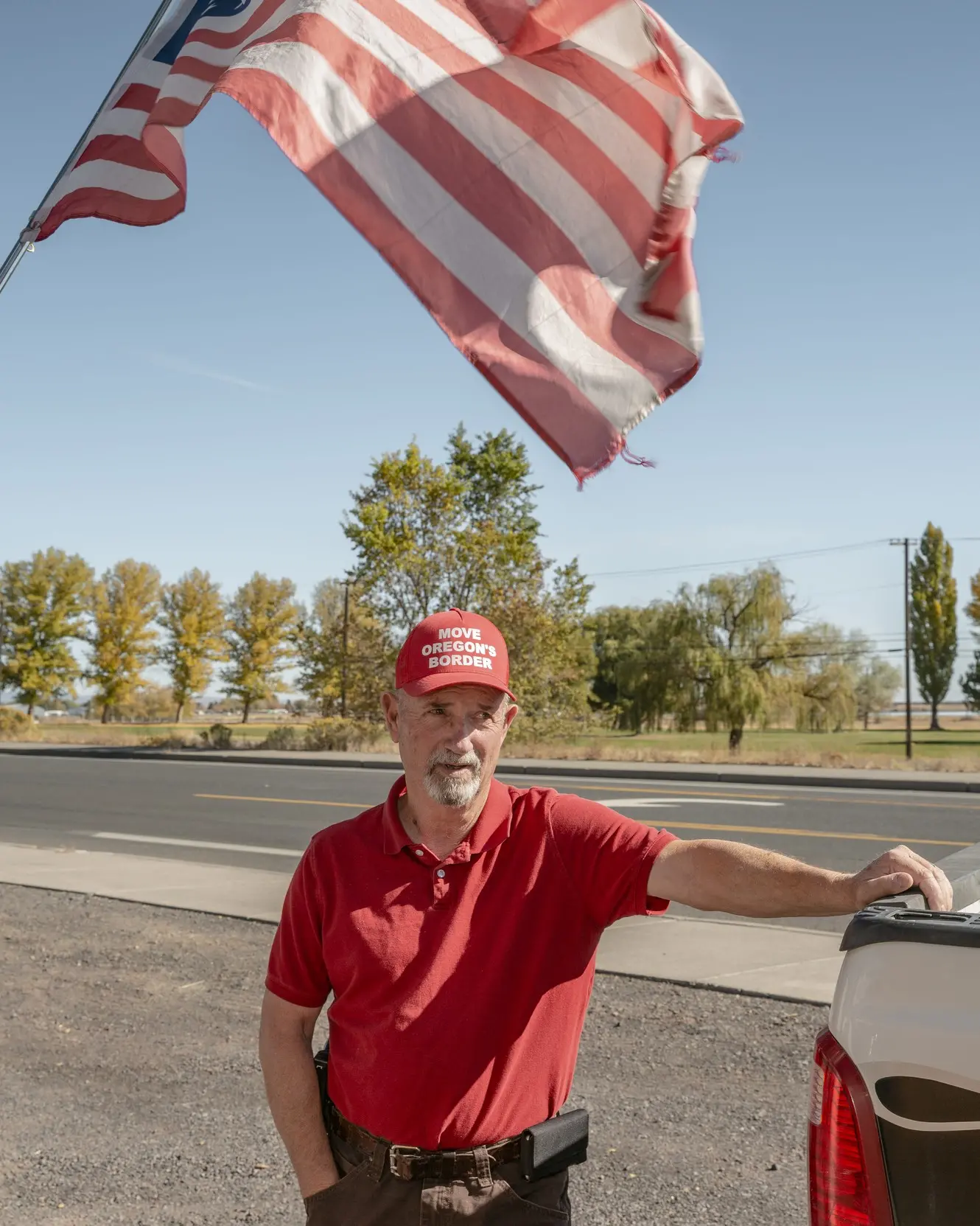 Man stands in front of truck and American flag wearing a red hat that says "Move Oregon's Border"