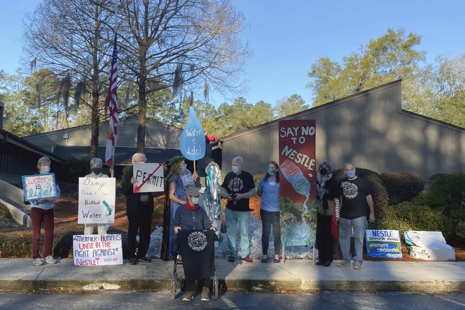 Protesters stand on a sidewalk outside of a wooden building. Their signs read: "water is life" , "deny permit for bottled water" and "say no to nestle"