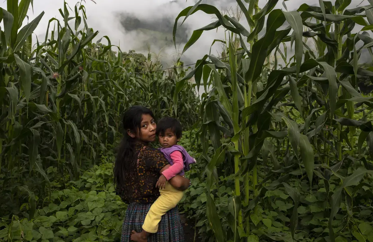a girl turns to face the camera while holding a young boy in a grassy field