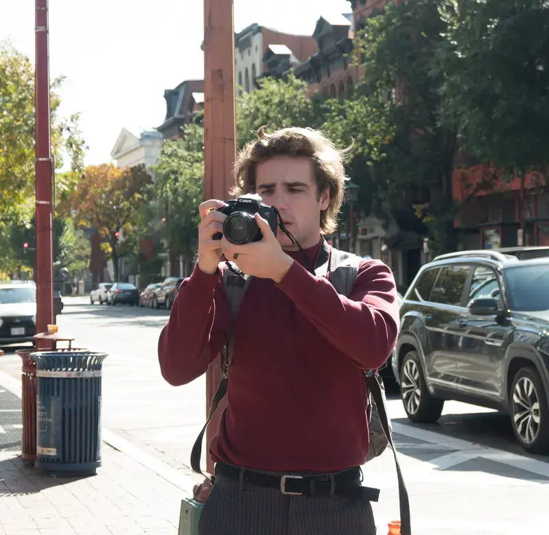 A person wearing a red shirt holds a camera up to their face in a city setting