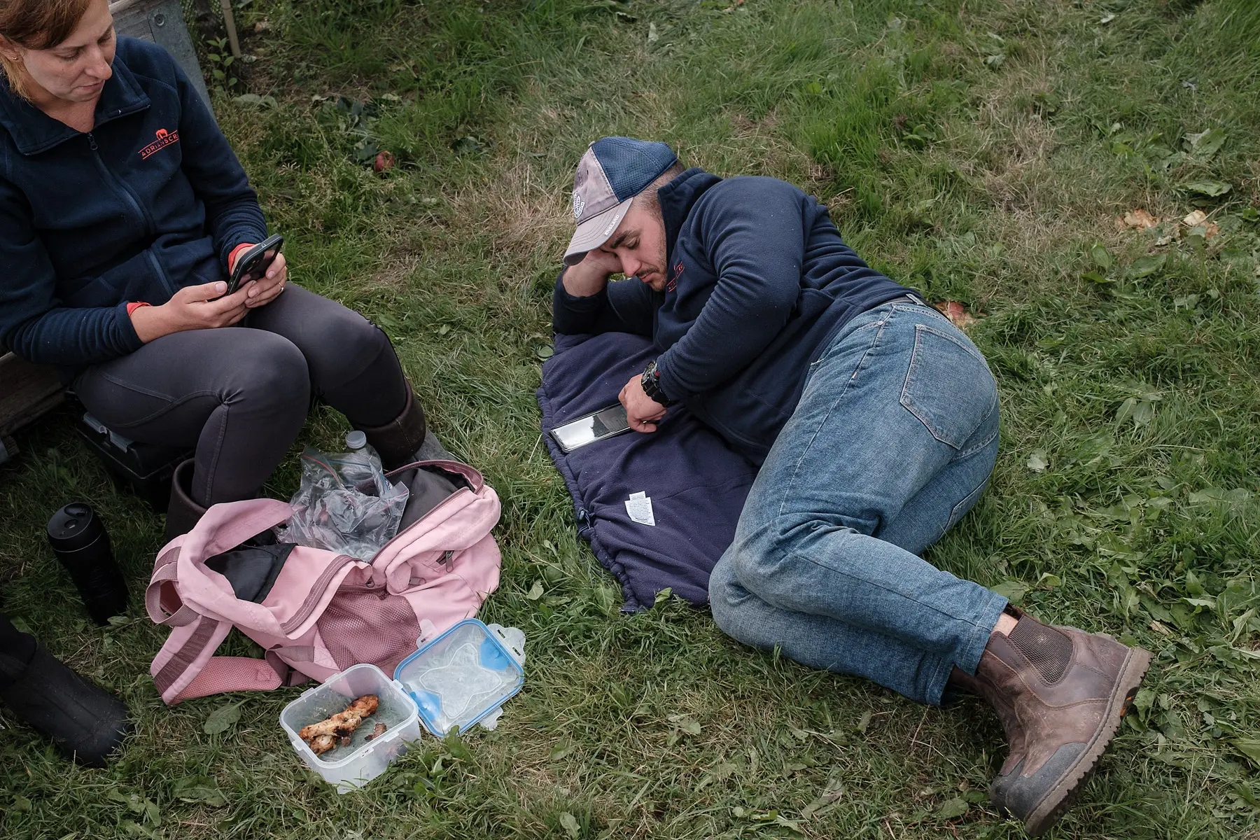 two farm workers rest in the grass with food, looking at their phones on their lunch break