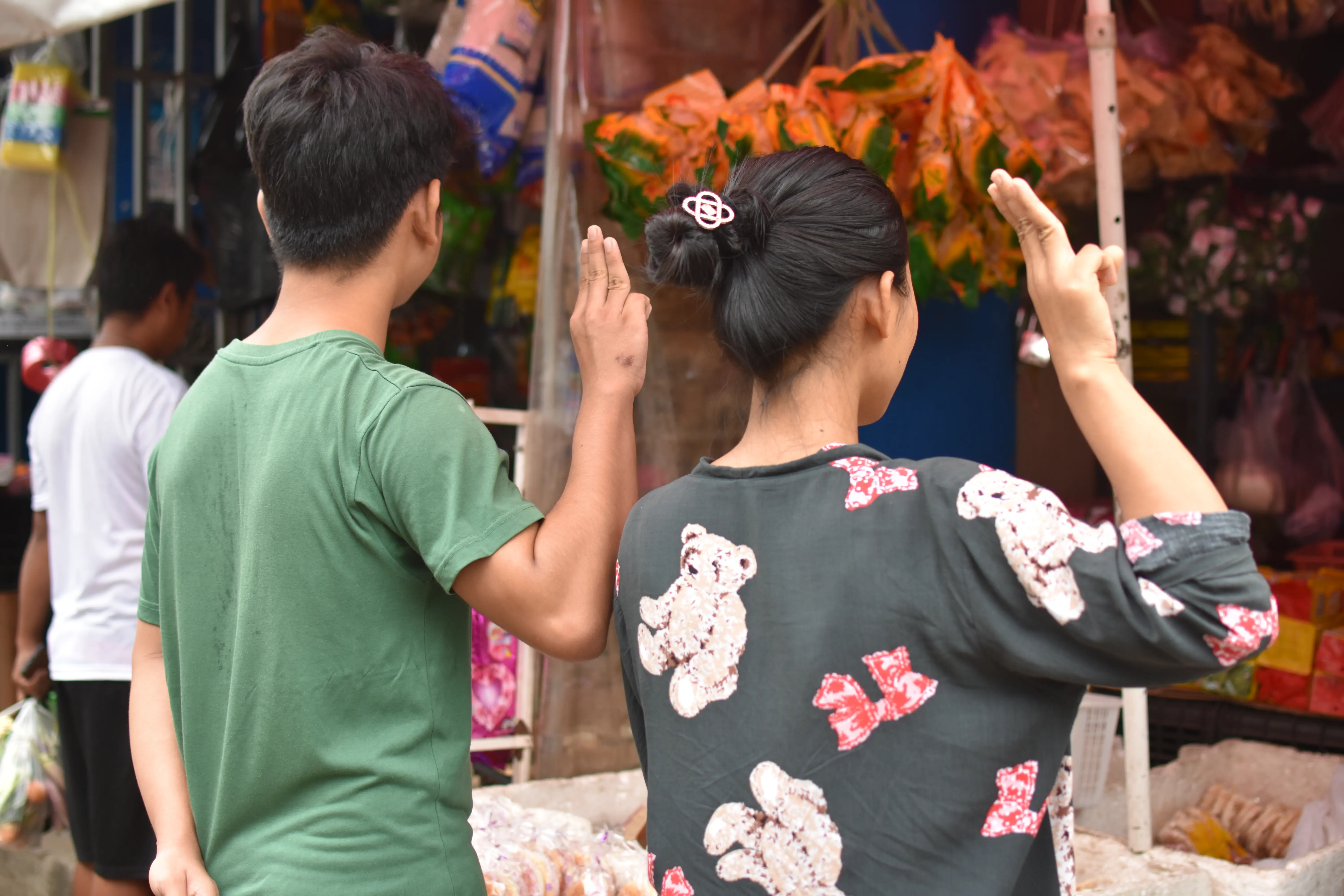 A man and woman hold up three fingers with their right hands in a market.