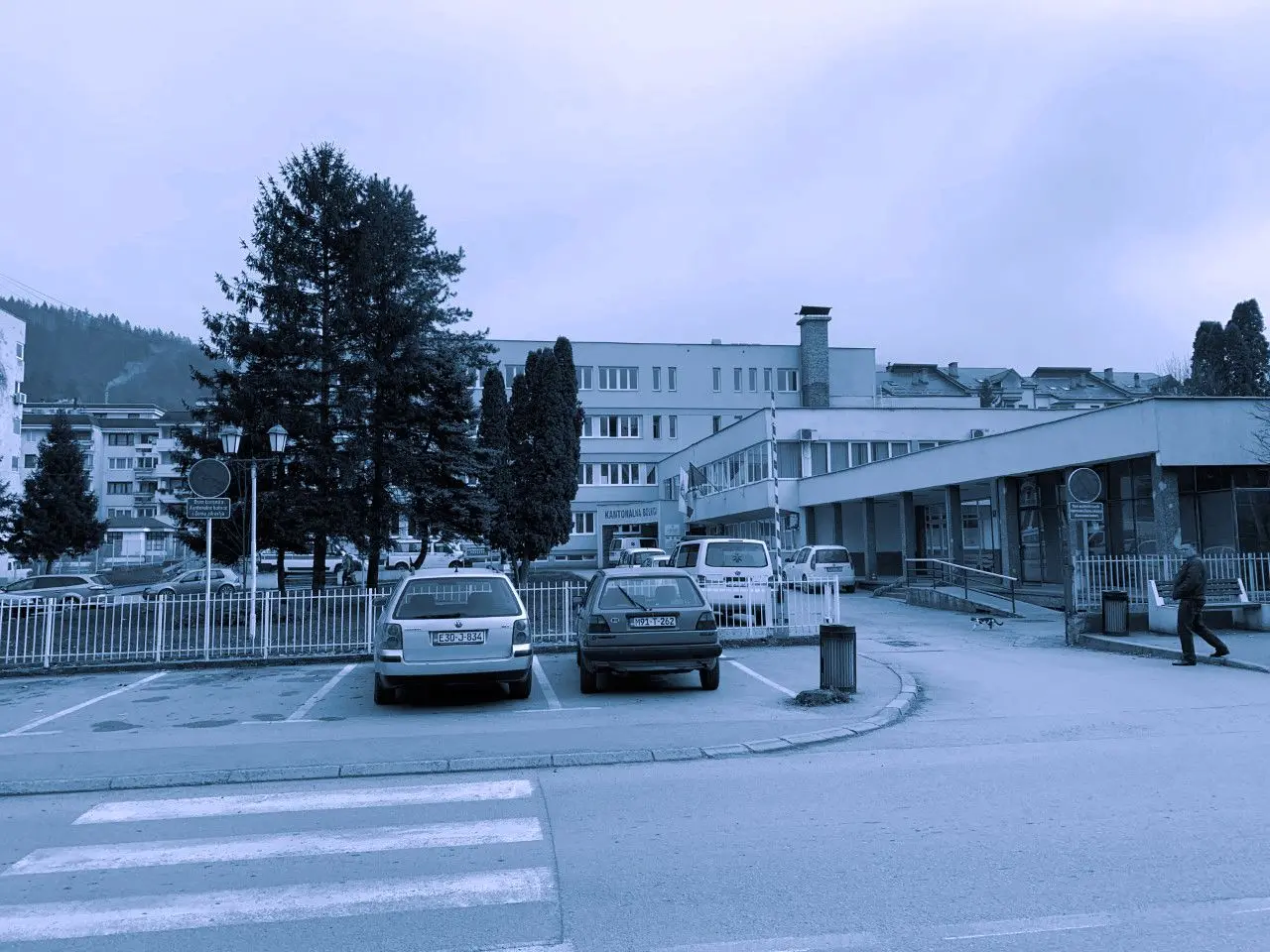 Blue-tinted image of a hospital with parking lot in front.