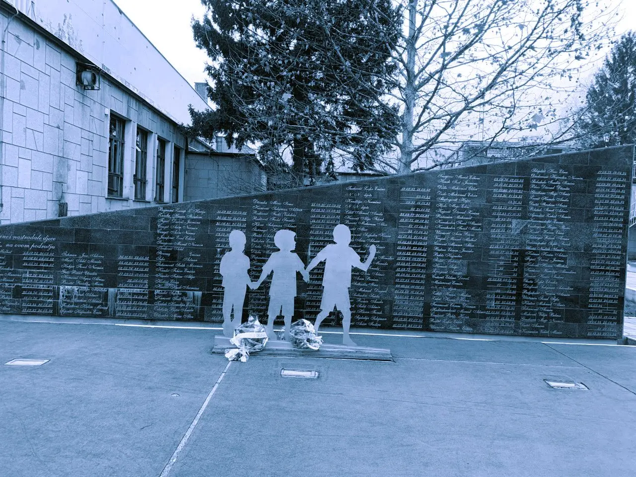 Blue-tinted photo of a memorial wall with names engraved on it. Silhouette statues of children stand in front of the wall.